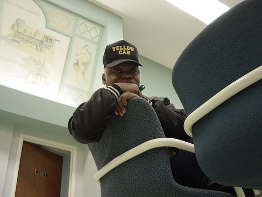 A cab driver in a Yellow Cab cap leaning on the back of a station chair
