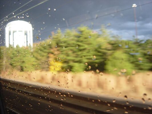 A white water tower and sunlit trees blurring past a rain-speckled train window, under dark clouds
