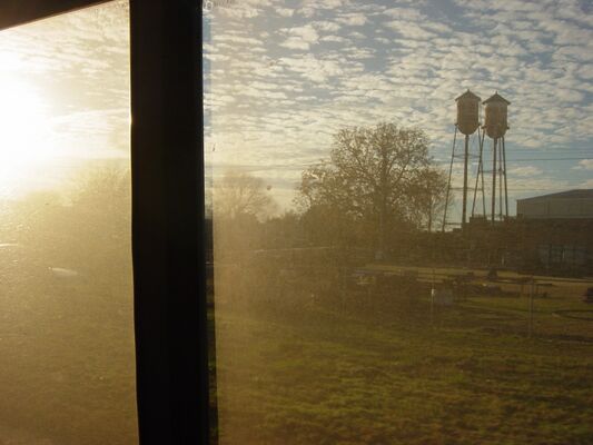 Two old water towers and a tree silhouetted against a hazy sunrise, seen past the edge of the train window