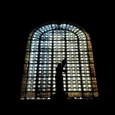 A figure silhouetted against a tall arched window of patterned glass in a dark church