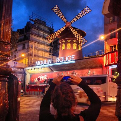 A woman photographing the glowing red windmill of the Moulin Rouge at dusk