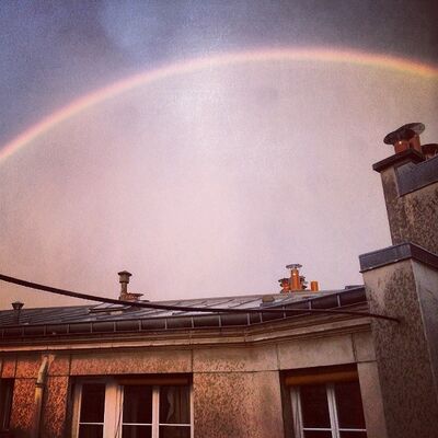 A faint rainbow arching over a Paris rooftop and its chimney pots