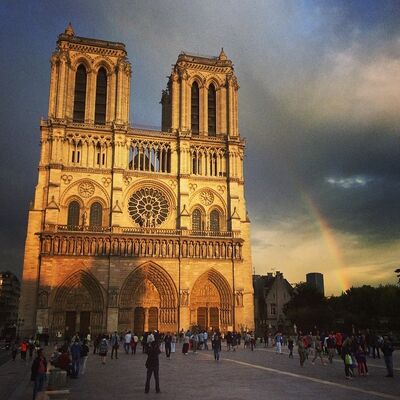 The front of Notre-Dame lit gold by the setting sun under a dark sky, with a rainbow beside it