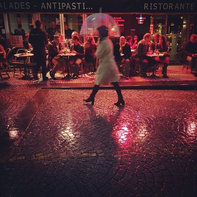 A woman with a clear umbrella walking past a busy café, its red lights reflected in wet cobblestones