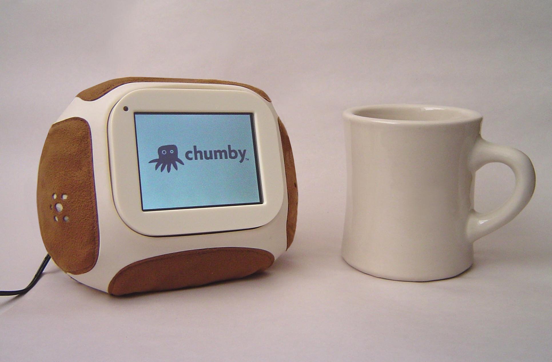 An original Chumby in its tan leather case, showing the Chumby logo on its screen, next to a white coffee mug