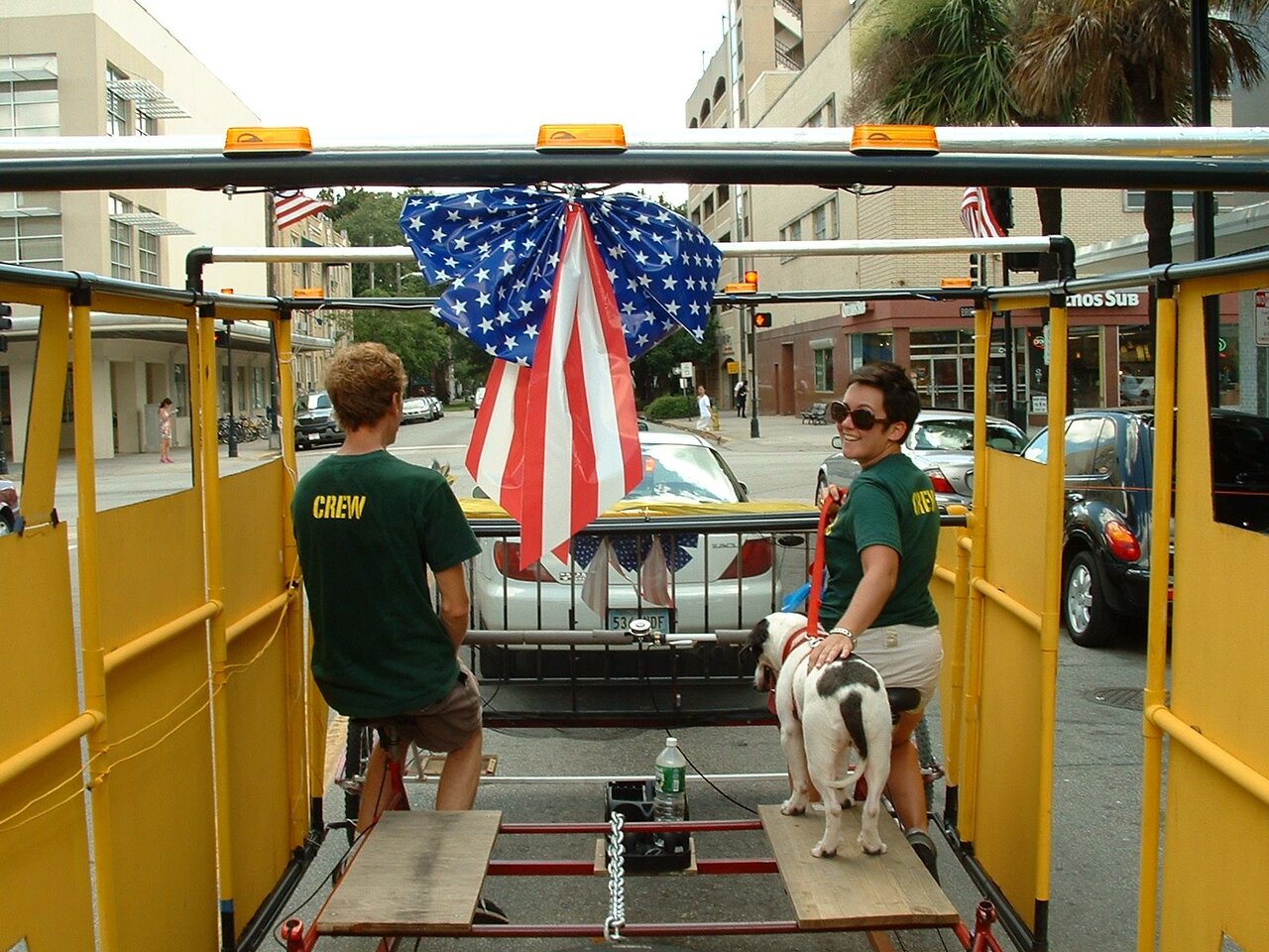 Two crew members in green Crew shirts pedaling the Green Hummer under a big stars-and-stripes bow, with a dog standing on one of the seats