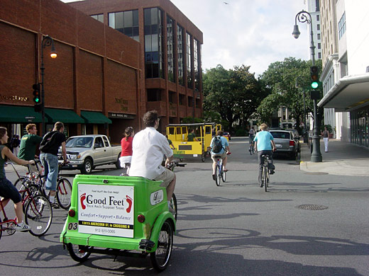 Cyclists and a green pedicab following the Green Hummer through a downtown intersection