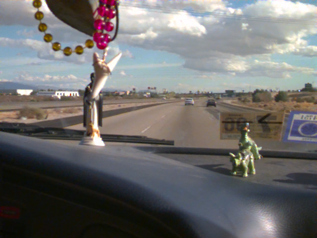 The highway through the windshield, with beads hanging from the mirror and a toy on the dashboard